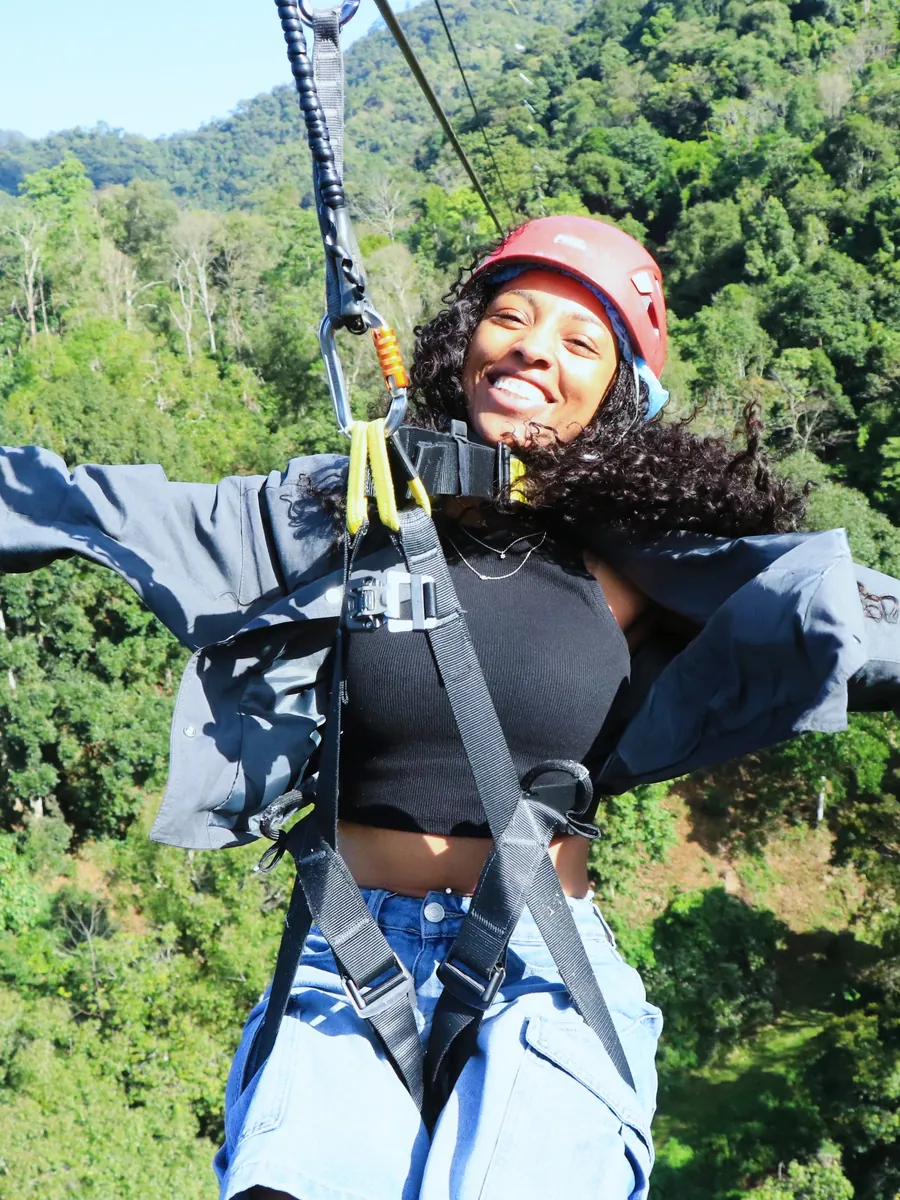 Kay in a red helmet and harness on a zipline above the jungle